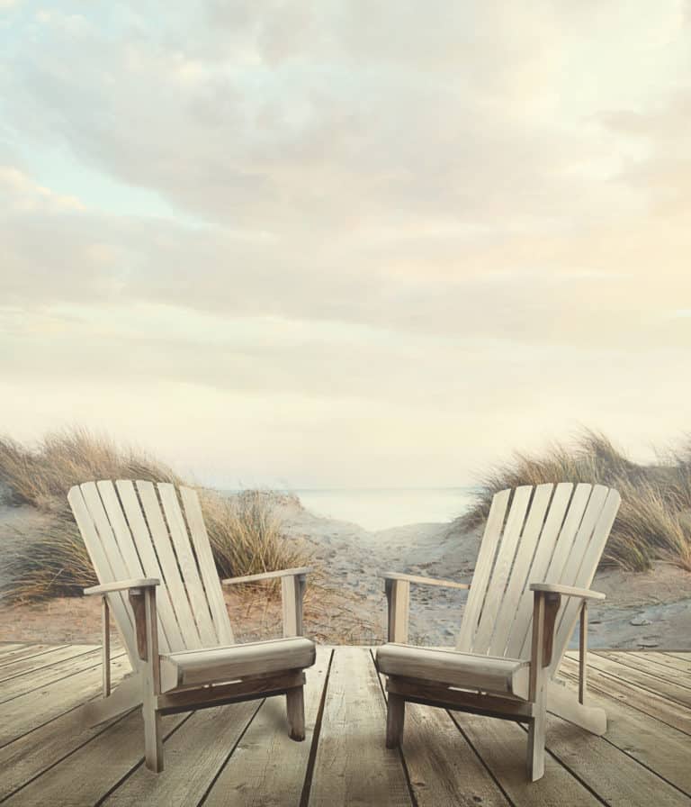 Two white Adirondack Chairs on a wooden deck at the beach.