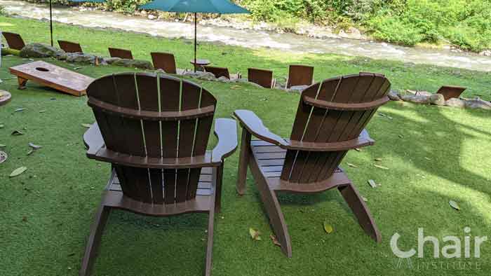 Two brown heavy-duty plastic Adirondack Chairs on the grass facing a rushing river.